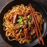 A close-up shot of Mongolian beef and noodles, served on a black plate, garnished with green onions and sesame seeds. card